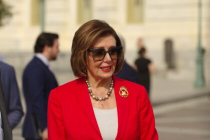 Nancy Pelosi smiling in a red blazer outside the U.S. Capitol building.