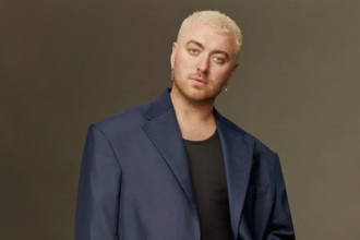 Sam Smith poses confidently in a sharp blue blazer against a dark background, looking directly at the camera with a poised expression.