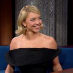 Sydney Sweeney sitting on The Late Show with Stephen Colbert sofa, animatedly telling the story of her skydiving adventure while smiling and gesturing.