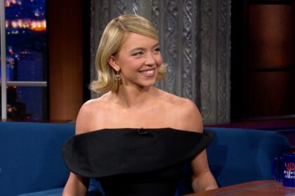 Sydney Sweeney sitting on The Late Show with Stephen Colbert sofa, animatedly telling the story of her skydiving adventure while smiling and gesturing.