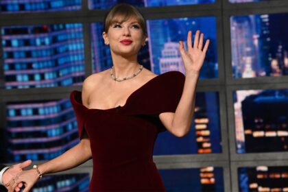 Taylor Swift waving to the crowd in a burgundy velvet David Koma minidress on the Late Show with Stephen Colbert stage, smiling confidently.